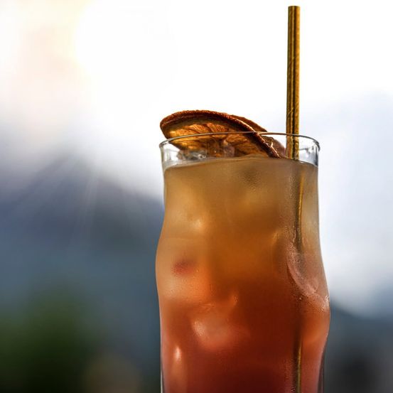 Ein hohes Cocktailglas mit einem goldenen Strohhalm, gefüllt mit einem mehrschichtigen Getränk, gekrönt von einer Orangenscheibe. Der Hintergrund ist unscharf mit einem hellen Licht.