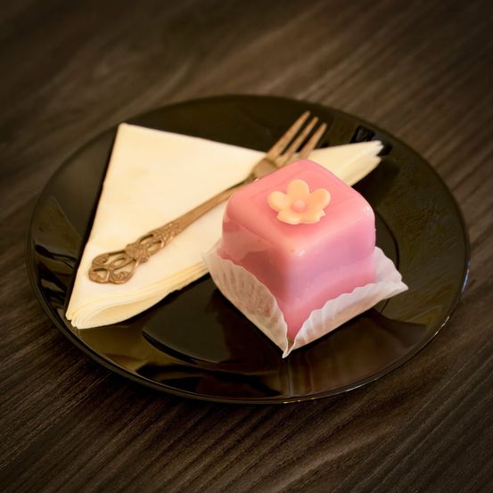 Ein rosafarbener quadratischer Kuchen mit einer Blume darauf steht auf einem schwarzen Teller mit einer weißen Serviette und einer goldenen Gabel.