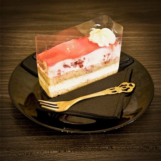 Ein Stück Erdbeer-Käsekuchen auf einem schwarzen Teller, gekrönt mit Schlagsahne und einer goldenen Gabel. Der Nachtisch ist in einer durchsichtigen Plastikhülle.