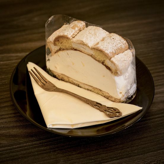 Ein Stück Kuchen mit einer Gabel auf einer Serviette, präsentiert auf einem schwarzen Teller mit einer durchsichtigen Plastikabdeckung. Der Kuchen hat eine cremige Füllung und ist mit Puderzucker bestreut.