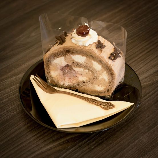 Ein Schokoladenkuchen mit Schlagsahne und einer Kirsche auf einem schwarzen Teller mit einer Gabel und einem Serviettentuch.
