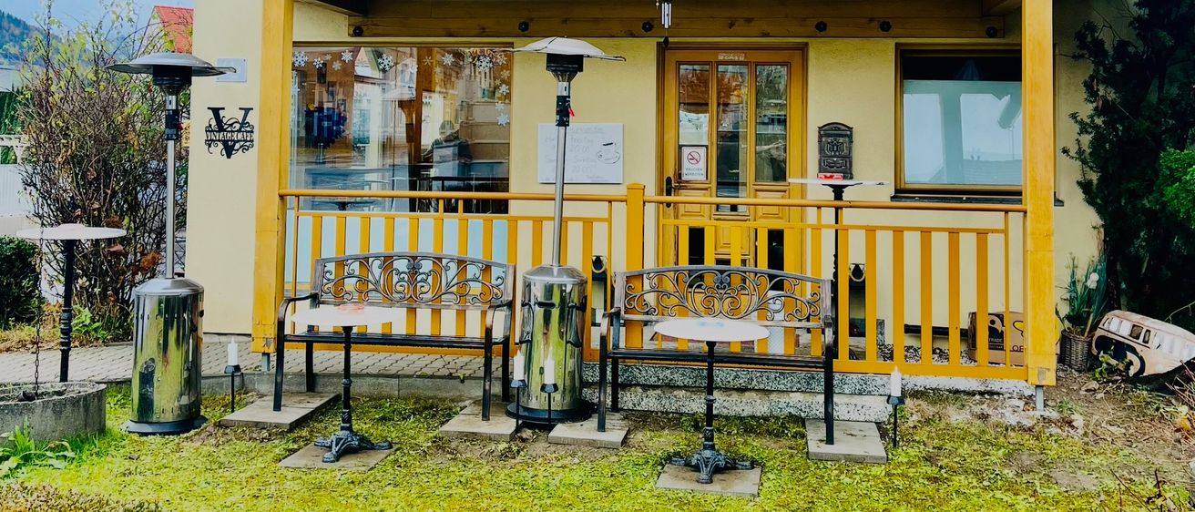 Ein kleines gelbes Haus mit rotem Dach hat zwei Bänke und Heizkörper draußen. Das Haus hat Glasfenster und eine Tür. Es gibt Bäume und Pflanzen in der Nähe.