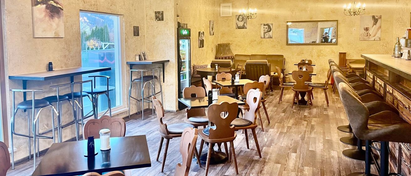 Ein Innenraum eines gemütlichen Restaurants mit Holzböden, herzförmigen Stühlen und einer Bartheke. Es gibt mehrere Tische und Stühle und ein Kühlschrank steht an der Wand.