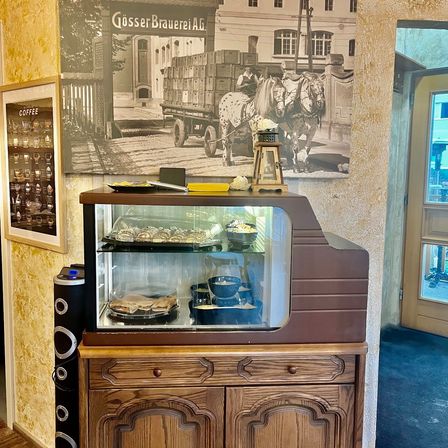 Ein Holzschrank mit einer Glasvitrine hält verschiedene Gebäckstücke. Dahinter befindet sich ein gerahmtes Bild mit Kaffeetassen an der Wand. Oben zeigt ein Foto Pferde, die einen Wagen ziehen.