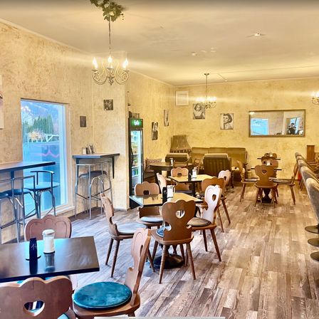 Das Innere eines Restaurants mit Holzboden, Holztischen und Stühlen, einem Glasfenster und einer Glastür. Die Decke hat eine Kronleuchter und Bilderrahmen. An den Wänden befinden sich Gemälde und ein Kühlschrank.