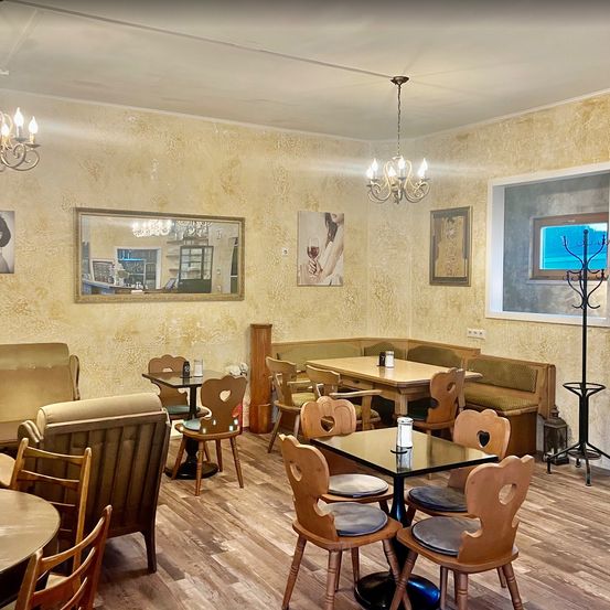 Ein elegantes Café-Interieur mit Holzböden, beigefarbenen Wänden und verschiedenen Möbeln wie Tischen, Stühlen und Sofas. Es gibt zwei Kronleuchter und gerahmte Bilder an der Wand.