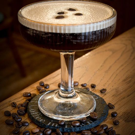 Ein Glas Kaffeecocktail mit schaumiger Spitze und verstreuten Kaffeebohnen auf einem Holztisch.