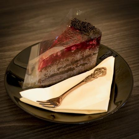 Ein Stück Schokoladenkuchen mit roter Beerenfüllung, in Plastik verpackt, und eine Gabel auf einem Serviettentuch auf einem schwarzen Teller.