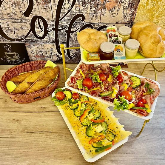Auf einem Holztisch sind verschiedene Sandwiches und Salate zu sehen, darunter ein Brotkorb und zwei Tabletts mit Fleisch, Gemüse und Brot.