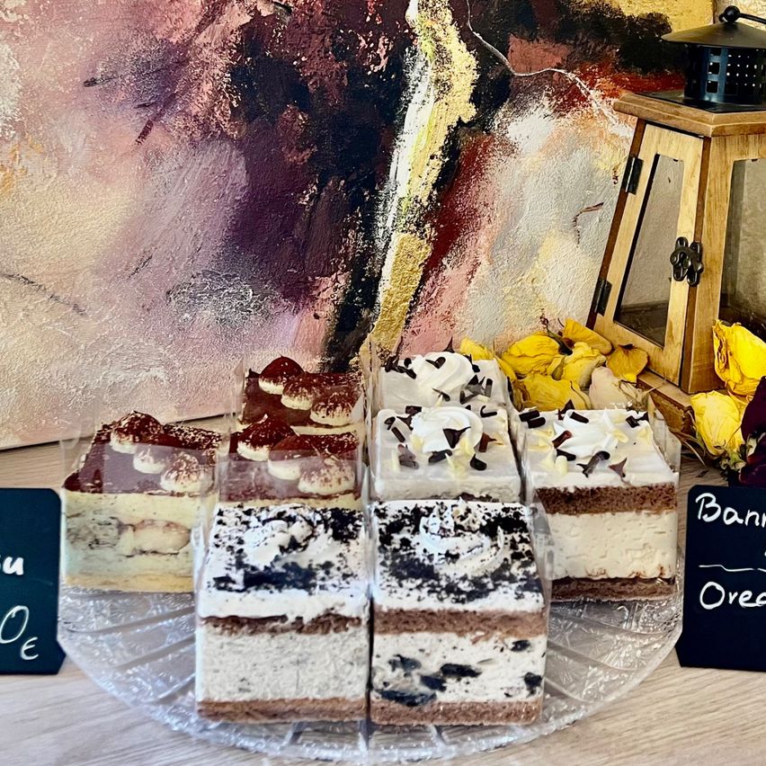 Auf einem Holztisch sind verschiedene Desserts in transparenten Boxen zu sehen, darunter ein Schokoladenkuchen mit weißer Creme und ein Vanillekuchen mit Schokolade. Dahinter ist ein großes abstraktes Gemälde und eine Holzlaterne zu erkennen.