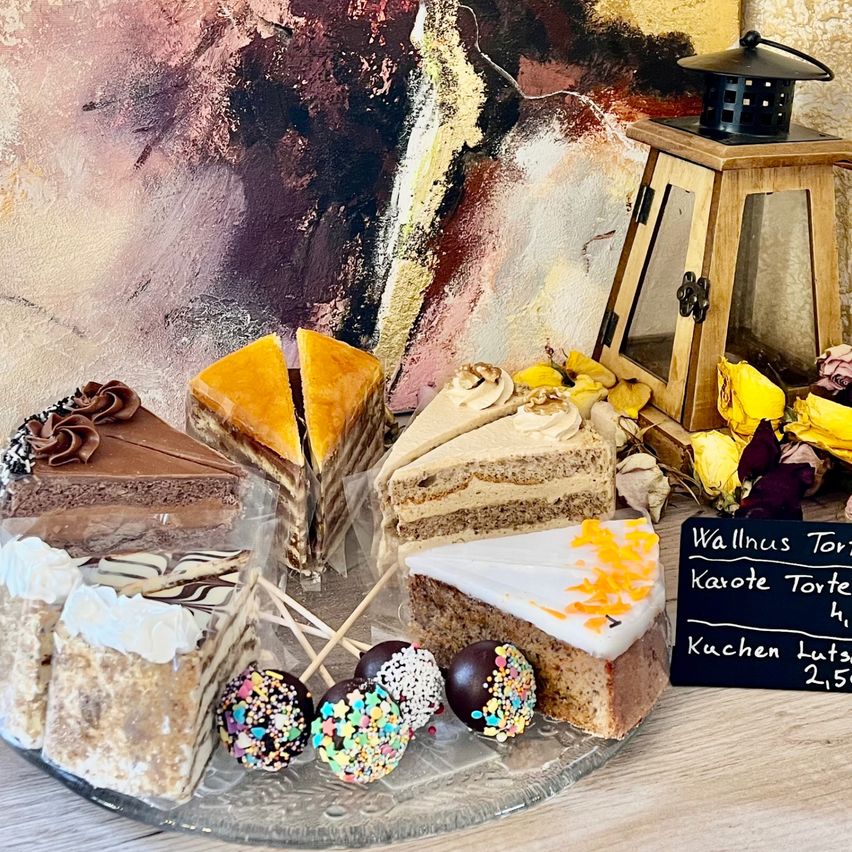Ein Tisch zeigt verschiedene Kuchen und Gebäck, darunter Schokoladen- und Karamellkuchenstücke, Karottenkuchen und einen Käsekuchen. Bunte Streusel schmücken Kuchenstangen. Dahinter steht eine Laterne und eine Wand mit einem abstrakten Gemälde. Ein schwarzes Schild mit Kuchennamen und Preisen.