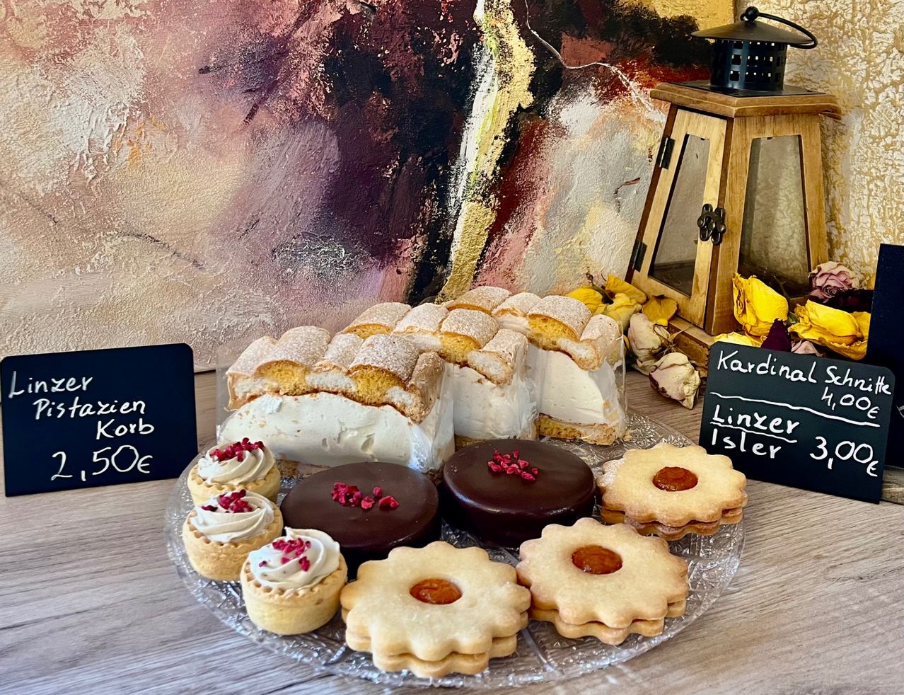 Ein Glasteller zeigt verschiedene Desserts, darunter Schichtkuchen, Schokoladenkuchen und Kekse, mit einem Schild, das 'Korb 50€' liest. Hinter dem Teller befindet sich eine Wand mit einer abstrakten Malerei und einer Laterne.