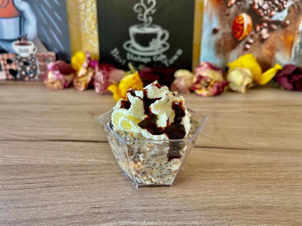 Ein Dessert mit Schlagsahne und Schokoladensauce in einem durchsichtigen Behälter auf einem Holztisch, mit Blumen und einem Café-Schild im Hintergrund.