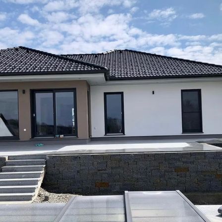 Ein modernes Haus mit einem Ziegeldach und Glaswänden, einer Steinmauer und einem Patio. Es gibt Treppen, die zum Eingang führen.