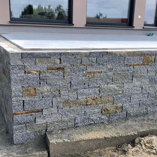 Ein Steinmauer wird vor einem Gebäude mit großen Fenstern und einer grauen Terrasse errichtet, mit Werkzeugen und Zement in der Nähe.