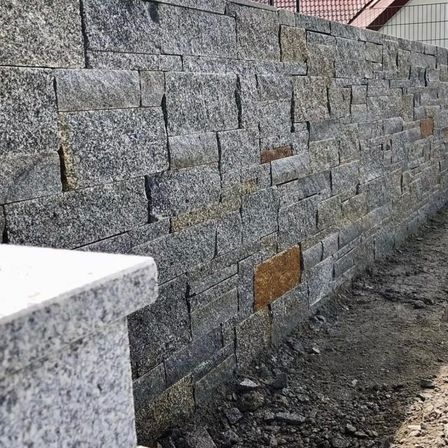 Eine hohe Granit-Stützmauer im Bau. Die Mauer besteht aus Granitblöcken mit verschiedenen Grau- und Brauntönen. Schmutz und kleine Steine sind um die Basis herum sichtbar.
