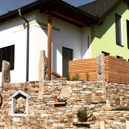 Ein modernes Haus mit weißen und grünen Wänden, einer Steinmauer und einem Holzbalkon, geschmückt mit Topfpflanzen und dekorativen Steinen.