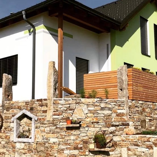 Ein modernes Haus mit weißen und grünen Wänden, einer Steinmauer und einem Holzbalkon, geschmückt mit Topfpflanzen und dekorativen Steinen.
