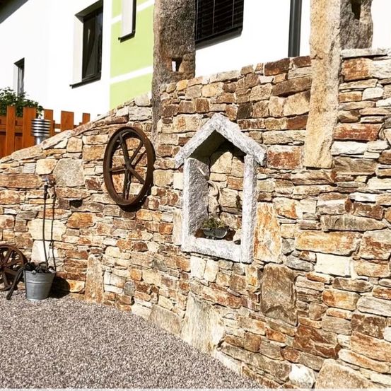 Eine Steinmauer mit einem Rad und einer kleinen Nische mit einer Topfpflanze. Die Mauer grenzt an ein Haus mit einem Holzzaun.