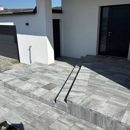 Das Äußere eines modernen Hauses mit zwei schwarzen Türen und einem grau gefliesten Boden. Das Haus hat eine weiße Wand und ein schwarzes Dach. Es gibt einen Schatten auf dem Boden.