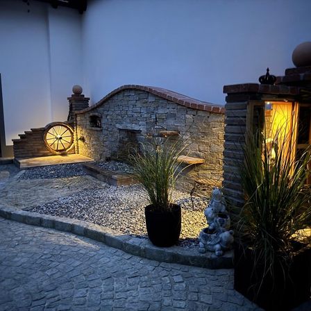 Eine Nachtansicht eines Außenbereichs mit einer Ziegelmauer, einem Brunnen und einem Rad. Es gibt zwei Pflanzen in schwarzen Töpfen und eine kleine Statue. Die Wand wird von einem gelben Licht beleuchtet.