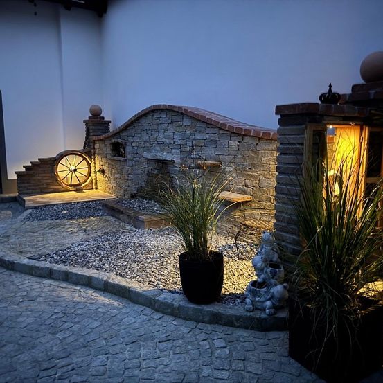 Eine Nachtansicht eines Außenbereichs mit einer Ziegelmauer, einem Brunnen und einem Rad. Es gibt zwei Pflanzen in schwarzen Töpfen und eine kleine Statue. Die Wand wird von einem gelben Licht beleuchtet.