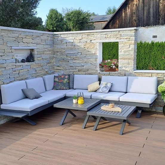 Eine Außenterrasse mit einem grauen Sofaset, zwei Tischen und einer Steinmauer mit Blumenarrangement.
