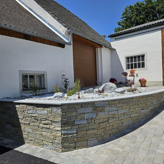 Außenansicht eines weißen Hauses mit einer braunen Garagentür und einer Steinmauer mit Pflanzen und Blumen.