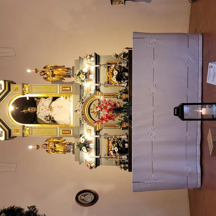 Ein Altar mit einer goldenen Statue, Blumen und einem Bilderrahmen mit einer Person darin. Ein weißer Tischläufer mit Mustern ist davor platziert.