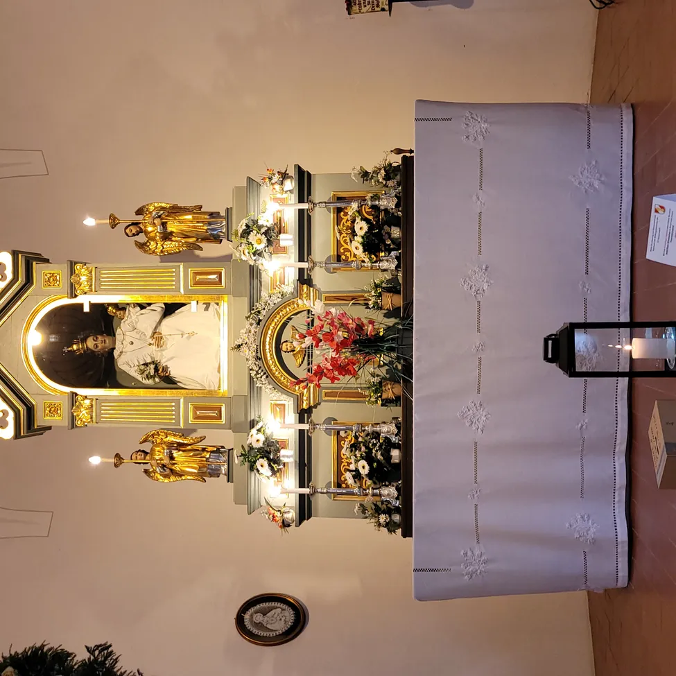 Ein Altar mit einer goldenen Statue, Blumen und einem Bilderrahmen mit einer Person darin. Ein weißer Tischläufer mit Mustern ist davor platziert.