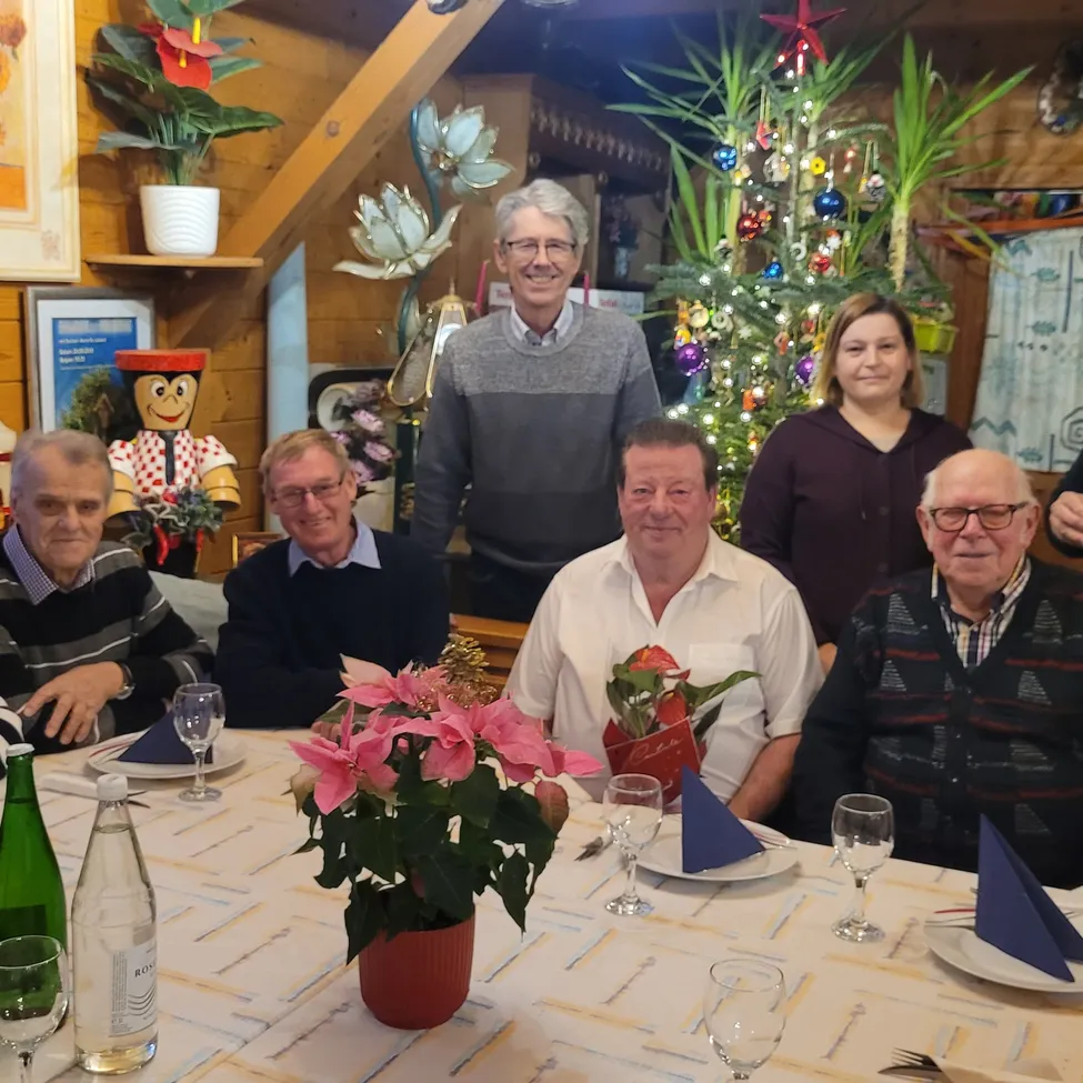 Eine Familie versammelt sich zu einem festlichen Abendessen um einen mit Blumen und einem Weihnachtsbaum geschmückten Tisch, wobei eine Holzwand und Dekorationen im Hintergrund zu sehen sind.