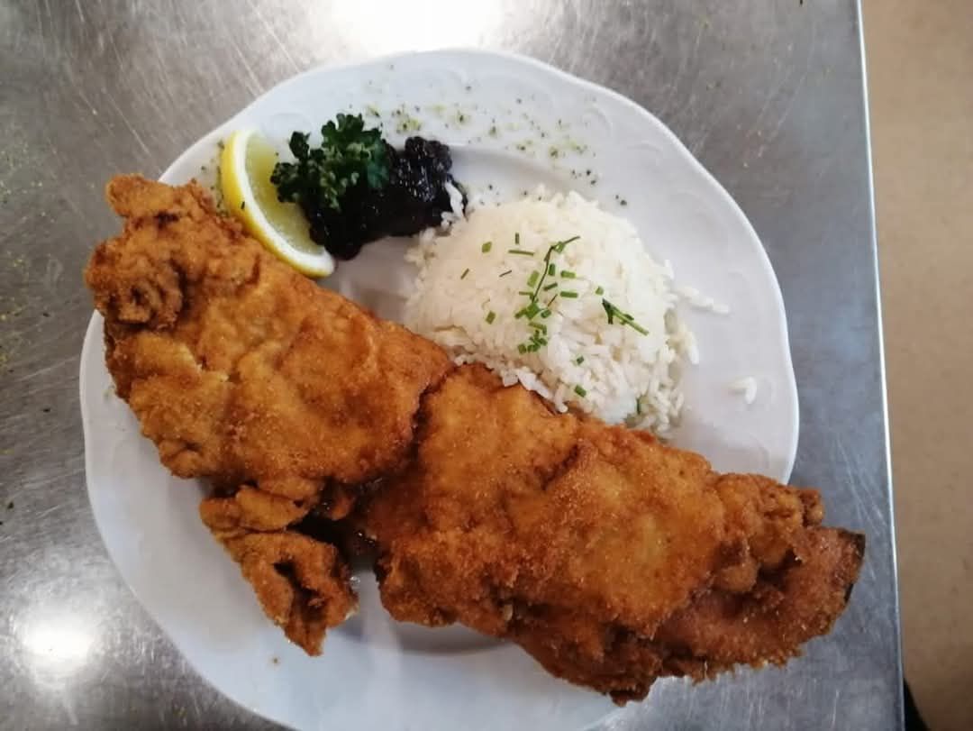 Ein Teller mit knusprigem gebratenem Fisch, serviert mit Reis, einer Zitronenscheibe und einer grünen Kräutergarnitur, auf einem weißen Teller auf einer Metalloberfläche.