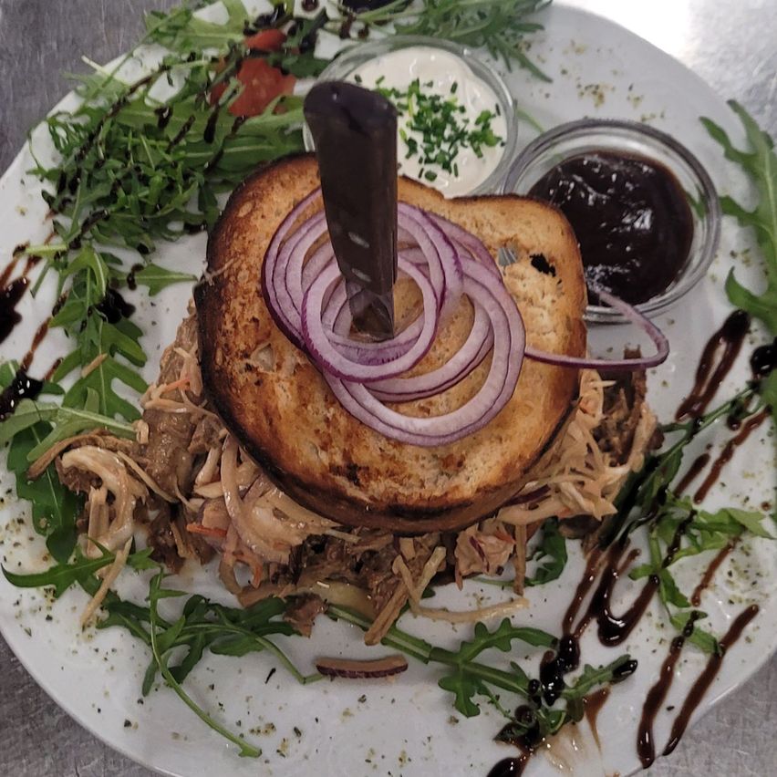 Ein Sandwich auf einem weißen Teller mit roten Zwiebeln, Rucola und einem Messer. Es gibt zwei Saucen in kleinen Schalen, eine mit einem grünen Kraut.