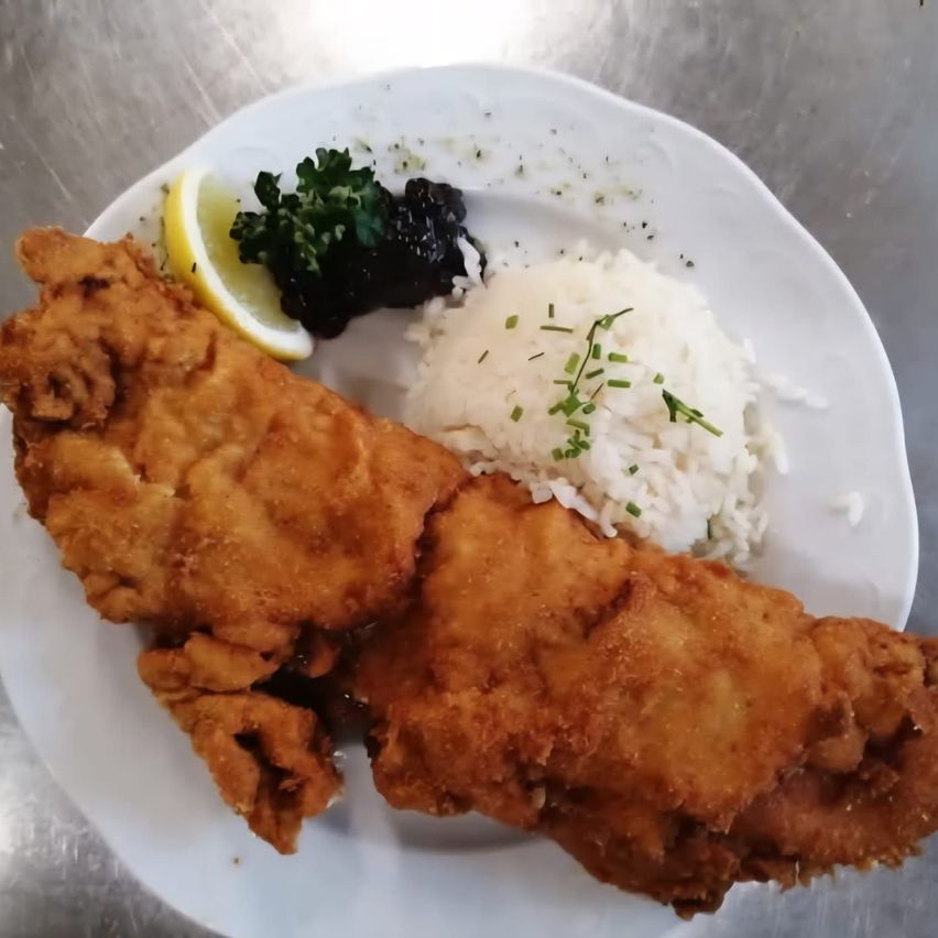 Ein weißer Teller enthält einen golden gebratenen Fisch, Reis und eine Zitronenscheibe. Auf dem Teller befindet sich auch eine kleine Portion schwarzer Sauce.