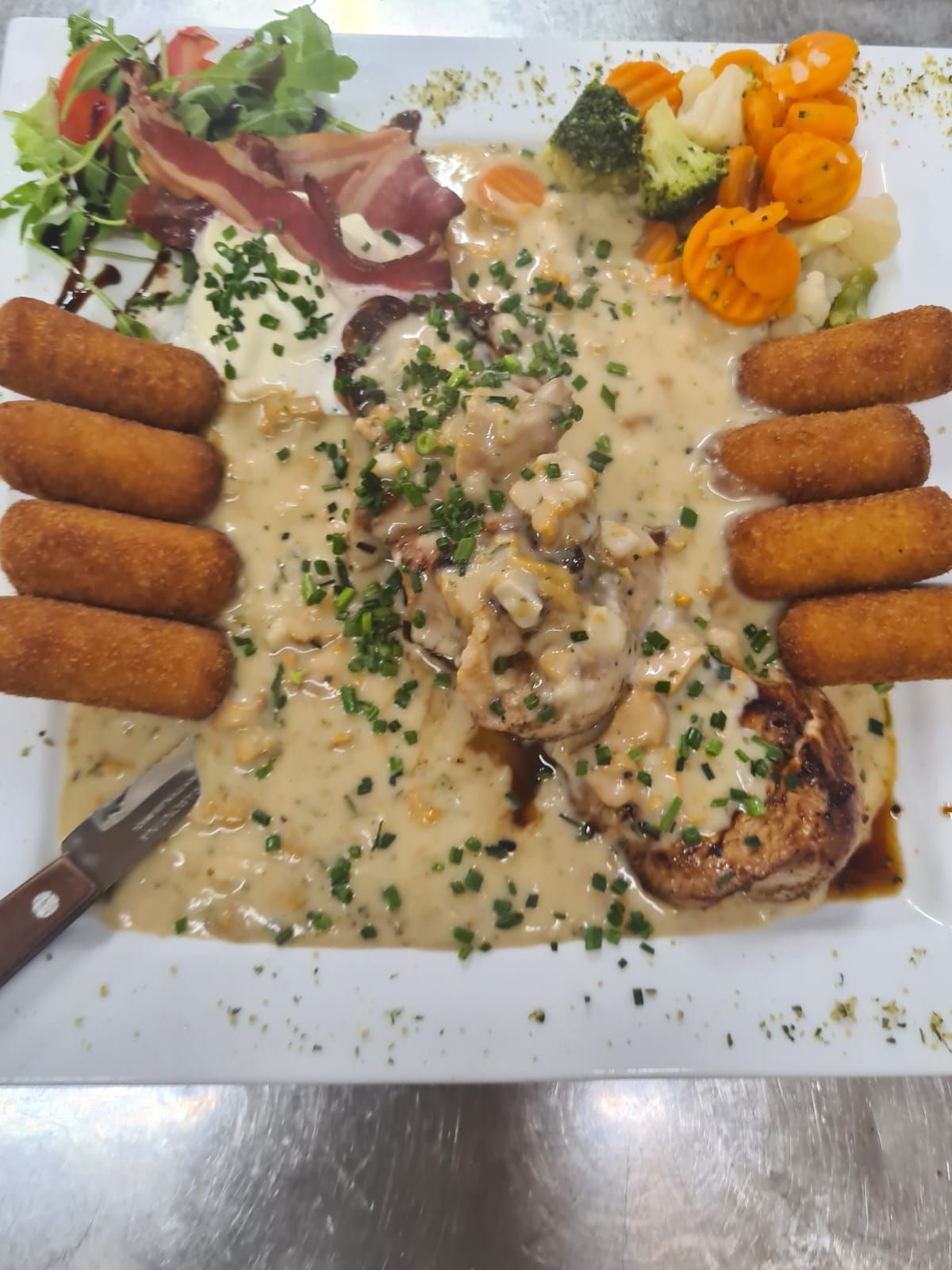 Ein Teller mit einer cremigen Sauce, Hähnchen, Gemüse und Fleisch, garniert mit grünen Kräutern und begleitet von einem Messer.