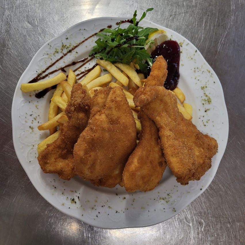 Ein Teller mit gebratenem Hähnchen und Pommes, garniert mit einem grünen Blattgemüse, einer Zitronenscheibe und einer roten Sauce auf einer metallischen Oberfläche.