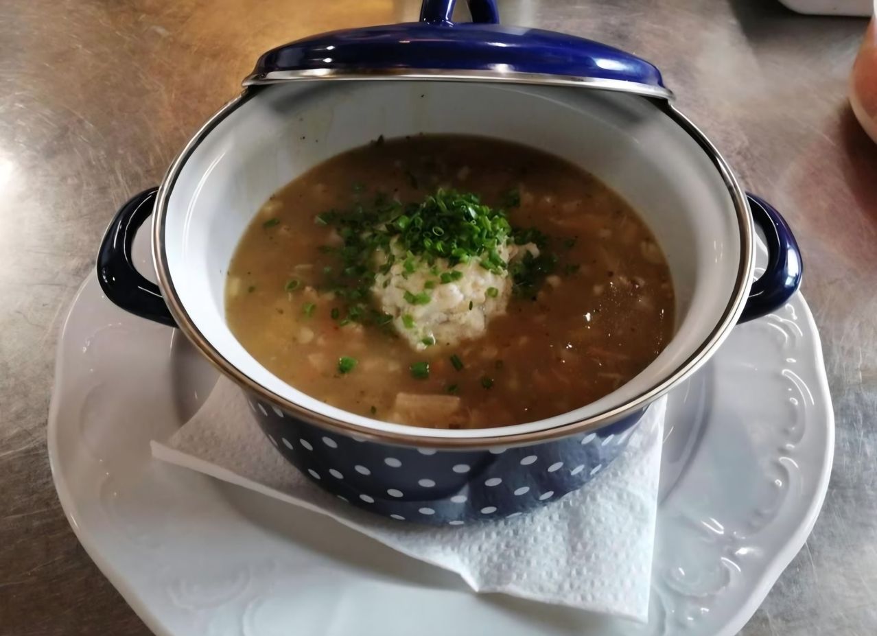 Eine Schüssel Suppe mit einem runden Stück Brot und grünen Kräutern darauf steht auf einem weißen Teller mit einem Serviettentuch auf einem Holztisch.