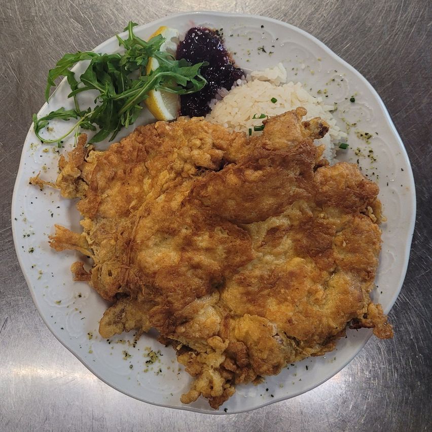 Ein Teller mit knusprigem gebratenem Hähnchen, Reis, Rucola und Marmelade mit einer Zitronenscheibe. Der Teller steht auf einer metallischen Oberfläche.
