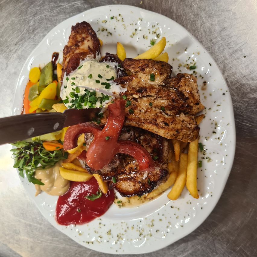 Ein Teller mit gegrilltem Hähnchen, Pommes und einer Auswahl an Gemüse, einschließlich einer Tomatenscheibe und einer Kette Sauce, mit einem Messer darauf.
