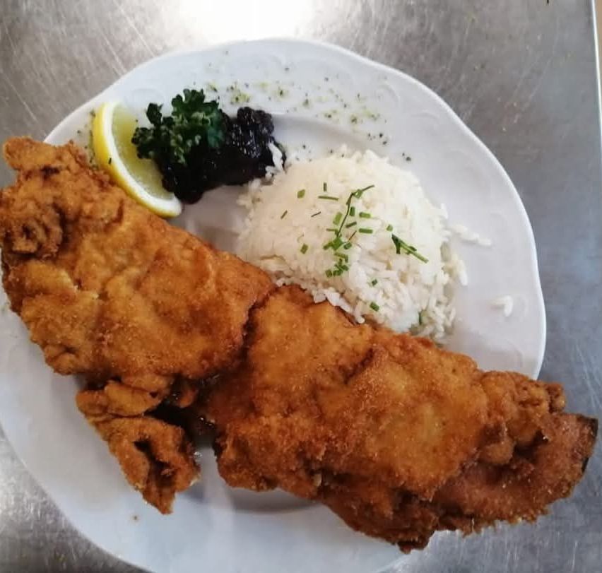 A white plate displays a meal consisting of two crispy fried fish fillets, white rice with green herbs, a lemon wedge, and a small black side dish.