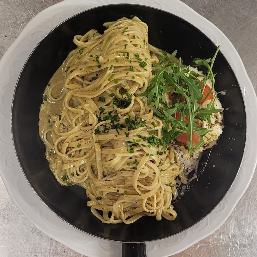 Ein schwarzer Teller mit Pasta, cremiger Soße und Rucola. Ein gebratenes Ei und gehackte Tomaten sind ebenfalls vorhanden.