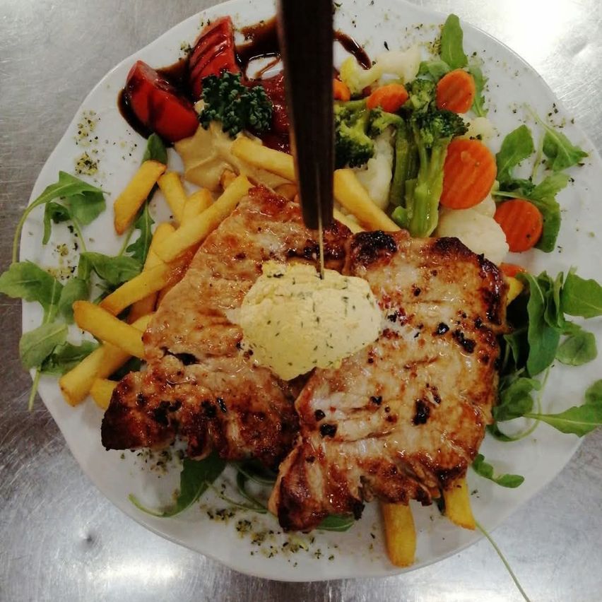A plate of grilled pork chops with a creamy sauce, accompanied by a variety of vegetables including broccoli, carrots, and arugula, and served with golden French fries.