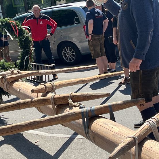 Eine Gruppe von Menschen steht hinter einem großen Holzstamm, der mit Seilen gefesselt ist. Ein Mann in einem roten Jackett steht vor einem silbernen Minivan.