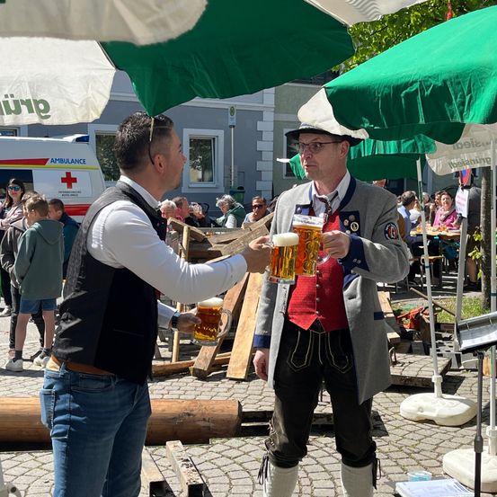 Zwei Männer in traditioneller Kleidung tauschen ein Glas Bier aus. Ein Mann trägt eine Weste und einen Hut, der andere eine rote Weste. Hinter ihnen sitzen mehrere Personen an Tischen unter Schirmen.