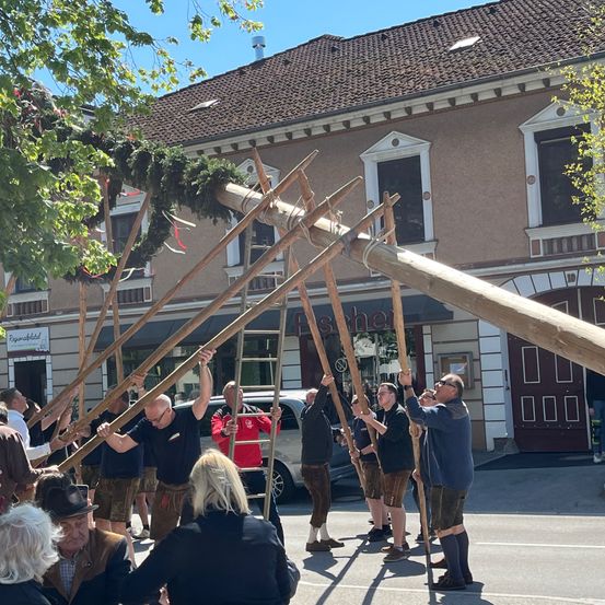 Men in traditioneller Kleidung halten lange Holzstangen vor einem Gebäude, mit einem Mann auf einer Leiter und einem Van im Hintergrund.
