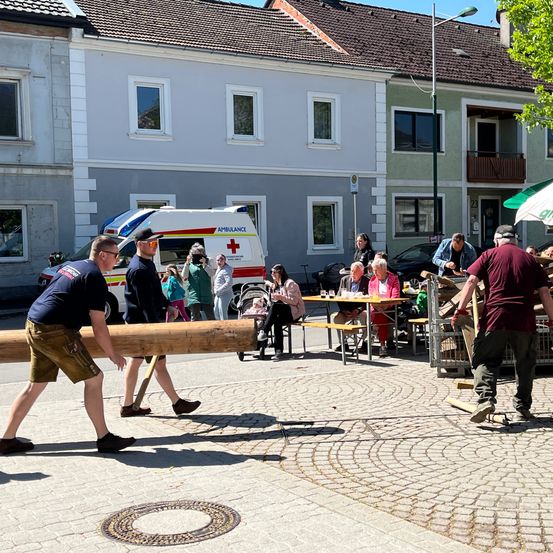 Zwei Männer tragen einen langen Baumstamm durch einen Stadtplatz, andere beobachten, darunter in einem Café. Dahinter ist ein Krankenwagen vor einem Gebäude geparkt.