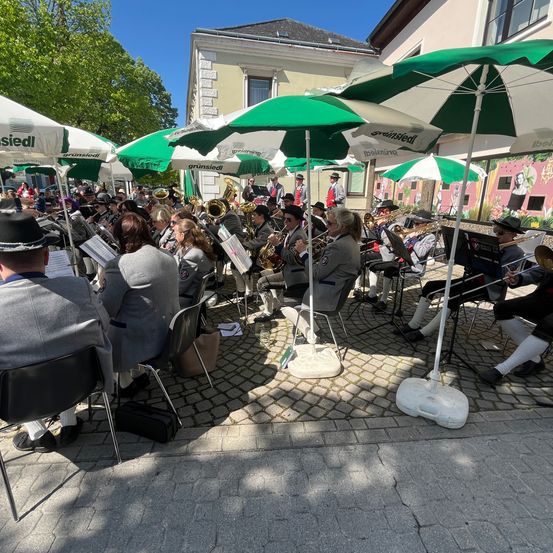 Ein Blasorchester spielt in einem von grünen Sonnenschirmen umgebenen Innenhof, mit Musikern auf Stühlen und Zuschauern, die zuschauen.