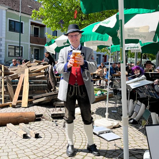 Ein Mann in traditioneller deutscher Kleidung, Hut und Bierglas in der Hand, steht auf einem gepflasterten Weg. Dahinter ist ein Stapel Holzplanken zu sehen, und es sitzen Leute an Tischen unter Schirmen. Eine Band spielt im Hintergrund, mit Instrumenten auf einem Tisch.