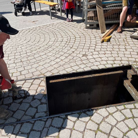 Eine Person benutzt ein Werkzeug, um einen Schachtdeckel auf einer Kopfsteinpflasterstraße zu öffnen, während andere in der Nähe sind.