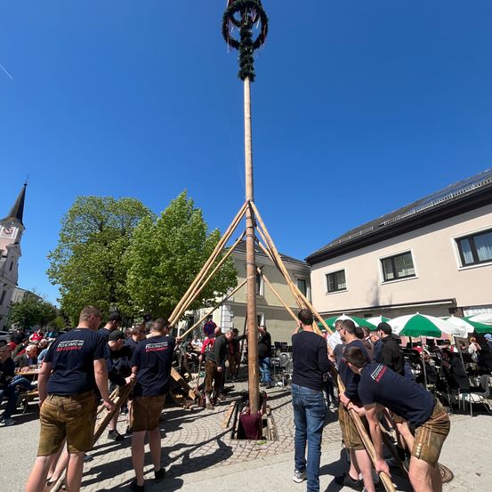 Eine Gruppe von Menschen hebt einen großen Holzpfahl mit Dekorationen auf der Oberseite an einem sonnigen Tag.